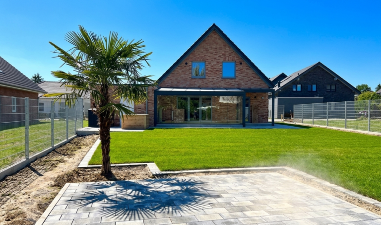 Modernes NRW-Einfamilienhaus mit Terrassenüberdachung und Glasschiebewand aus Aluminium, Sommerfoto mit Palme und Rasen